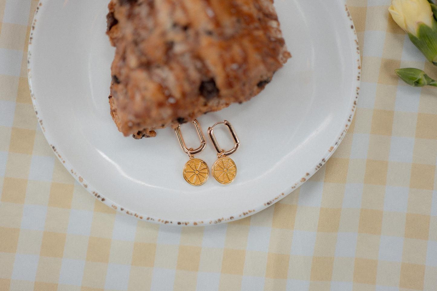 Gold earrings on a white plate with a piece of cake and flowers on a checkered tablecloth