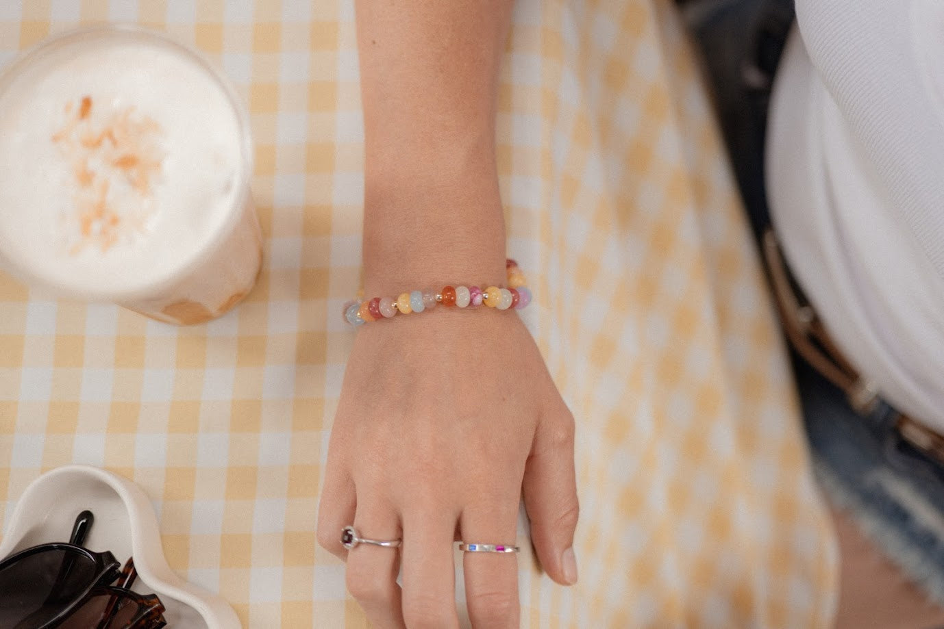 Hand wearing a colorful beaded bracelet on a checkered tablecloth with a candle and glasses.