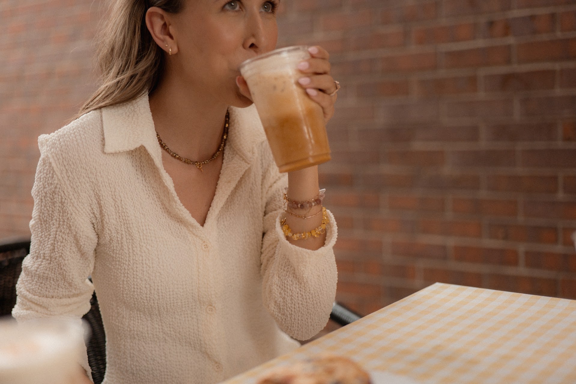 Woman drinking from a glass of iced coffee in front of a brick wall.